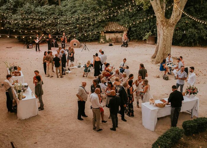 prise de vue en plongée sur la terrasse du Moulin et la fête d'été qui s'y trouve, les guirlandes scintillant et protégée par les platanes, on voit les invités se retrouver auprès des bars et buffets ou s'amuser sur la terrasse.