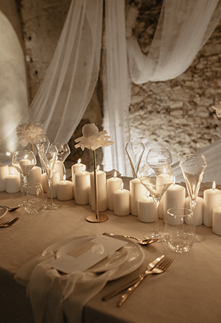 Prise de vue semi resserrée de la même table blanche tout en drapé et bougies blanches de nuit éclairée uniquement par les centaines de bougies en son centre.