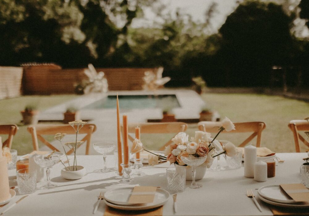 prise de vue en plan semi large avec un flou d'arrière plan sur une table de mariage blanche et jaune en bord de piscine avec un gâteau oranger, des bougies et des chandelles jaunes et des fleurs dans les mêmes tons.