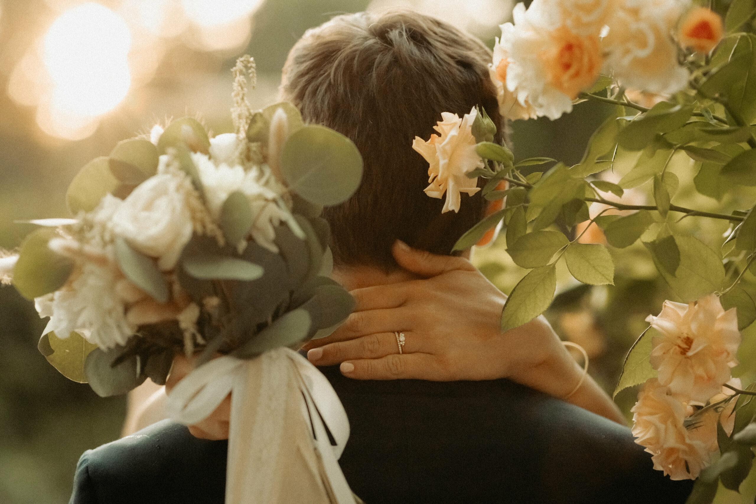 plan resserré des mariés de dos, on voix les mains de la mariés enlaçant le coup de son mari, le bouquet toujours en main, le tout au bord un rosier en fleurs