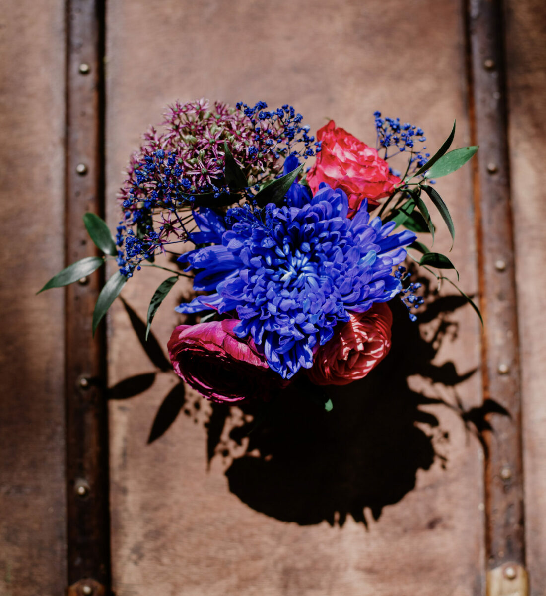 vue de haut sur un bouquet rouge et bleu, posé sur une valise vintage en cuir marroné et polie par le temps