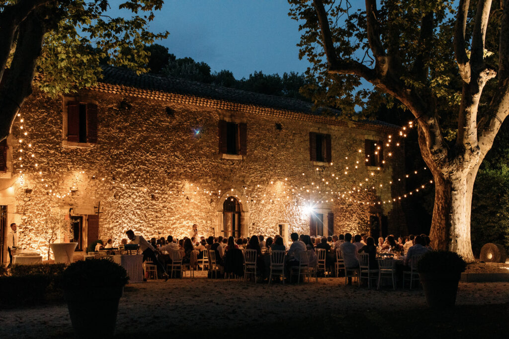 plan large d'une fête au Moulin, vu de l'exterieur, de nuit, éclairée par les guirlandes lumineuses entre les platanes et le toit