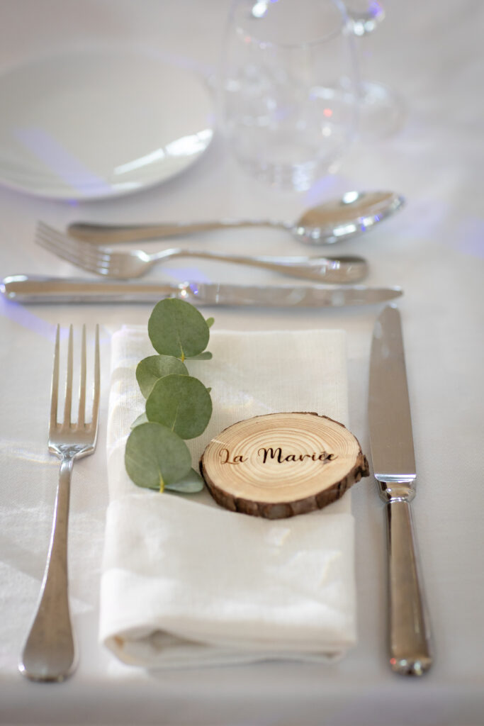 Plan resseré d'une place sur une table de mariage avec les couverts, la serviette et au dessus un goddie de bois au nom de l'invité.